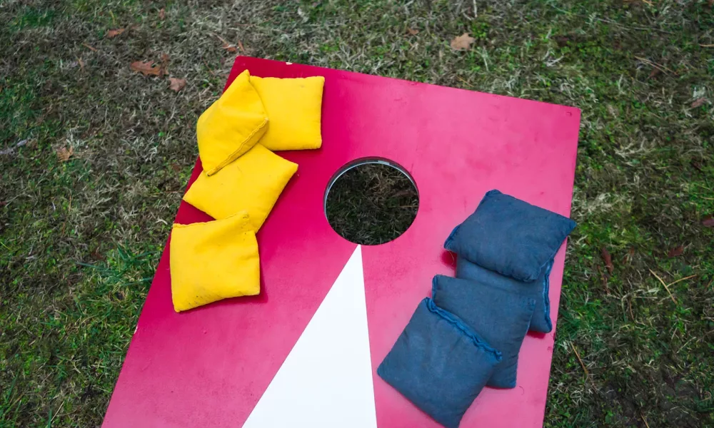 Above view of cornhole board with blue and yellow beanbags on green grass.